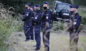 Policjanci w pełnym umundurowaniu, w tym w maskach, stoją w gotowości. W tle widać radiowóz. Sytuacja wygląda na stan wyjątkowy.