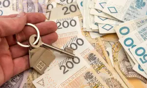 Dłoń trzyma klucz z breloczkiem w kształcie domu na tle polskich banknotów. Symbolizuje to możliwość zakupu nieruchomości lub dziedziczenia, co wiąże się z kwestią zachowku.