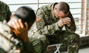 Żołnierze w mundurach siedzą, jeden z nich z rękami na twarzy, co może symbolizować walkę z PTSD.