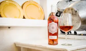 Un bicchiere e una bottiglia di negroamaro rosato Calitro su un tavolo bianco, con piatti gialli e pentole sullo sfondo.