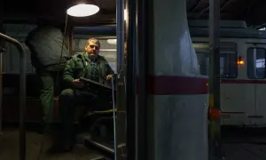 Mężczyzna w zielonym kombinezonie siedzi w kabinie tramwaju, trzymając panel sterowania. To fotografia miejska z wnętrza zajezdni.