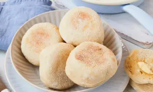Ein Teller mit vier goldbraunen englischen Muffins, daneben ein halber, mit Butter bestrichener Muffin. Ein Spiegelei ist im Hintergrund zu sehen.