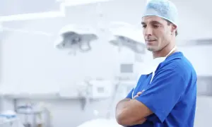 Chirurg w niebieskim kitlu i czepku stoi ze skrzyżowanymi rękami w sali operacyjnej.