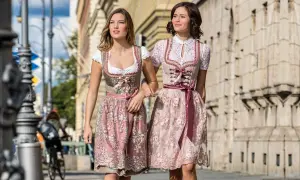 Zwei Frauen in Dirndln ohne Schnürung spazieren. Die Kleider sind in Rosa- und Beigetönen gehalten und mit zarten Stickereien verziert.