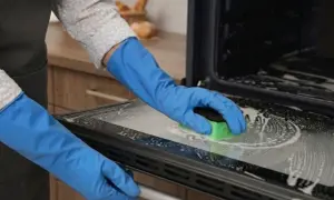 Ręce w niebieskich rękawiczkach czyszczą gąbką przypalone szkło hartowane piekarnika, usuwając zabrudzenia.