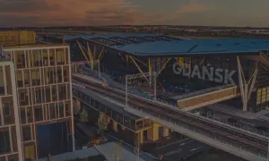 Nowoczesny budynek lotniska Gdańsk z napisem "GDAŃSK" na fasadzie, widok z góry. Widać tory kolejowe i perony.