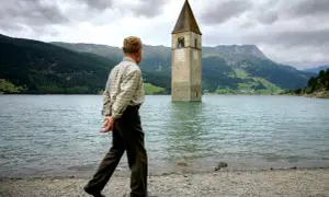 Ein Mann spaziert am Ufer eines Sees, auf dem ein Kirchturm aus dem Wasser ragt. Die Kirche im Stausee ist ein eindrucksvolles Zeugnis der Vergangenheit.