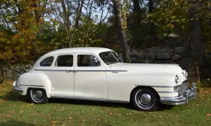 En vit Chrysler New Yorker från 1940-talet står parkerad på en gräsmatta med höstlöv.