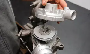 Dłoń trzyma część turbosprężarki nad jej głównym korpusem z widocznym wirnikiem.