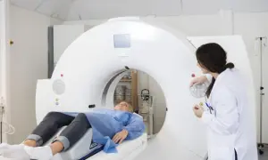 Pacjentka leży na stole tomografu, przygotowana do badania. Lekarka w białym fartuchu obsługuje aparat, który wykonuje angiografię.