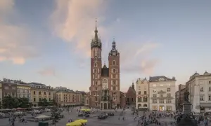 Krakowski Rynek Główny z majestatyczną Bazyliką Mariacką w tle. Ludzie spacerują, a nad nimi pastelowe chmury.