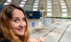 Une jeune femme souriante explore une piscine tournesol abandonnée, son dôme blanc orné de fenêtres rondes.