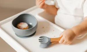 Małe rączki chwytają sztućce, gdy dziecko samo je. Miseczka z jedzeniem i okruszki na tacce.