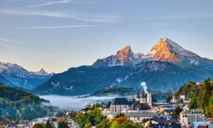 Morgensonne beleuchtet den spitzen Gipfel des kleinen Watzmanns über einem nebelverhangenen Tal und einer malerischen Stadt mit Kirchtürmen.