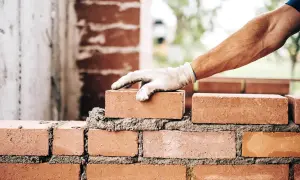 Dłoń w rękawicy kładzie cegłę na mokrym cemencie, budując ścianę.
