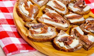 Kromki chleba z masłem i filecikami anchois ułożone na drewnianej desce, obok czerwono-biała serwetka.