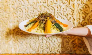 Un plat de couscous algérien, garni de légumes colorés et d'une viande savoureuse, est présenté sur un fond orné.
