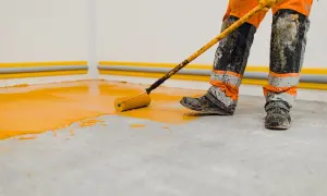 Un ouvrier en combinaison orange et noire utilise un rouleau pour peindre le sol d'un garage en jaune vif.