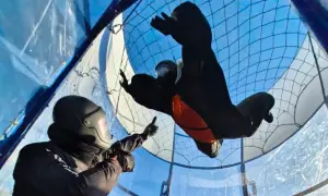 Dwóch śmiałków w kombinezonach i kaskach doświadcza lotu w tunelu aerodynamicznym, unosząc się w powietrzu.
