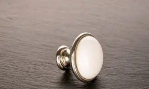 Elegancka kołatka meblowa z białym, ceramicznym elementem i metalową ramką, leżąca na ciemnym, kamiennym tle.