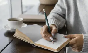 Mano escribiendo en un diario, un ritual de autocuidado. El café humeante acompaña este momento de introspección, una forma de escribir un diario como terapia.