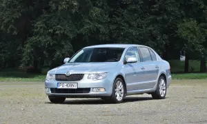 Jasnoniebieska Skoda Superb 2 z nowymi żarówkami, zaparkowana na placu przed drzewami.