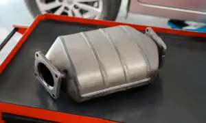 Metalowy katalizator samochodowy leżący na czarnej powierzchni z czerwonymi elementami.