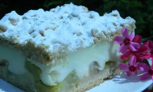 Pyszne ciasto z rabarbarem i budyniem, posypane cukrem pudrem. Obok kuszą różowe kwiaty.