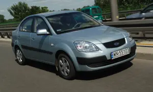 Błękitne światła Kia Rio na drodze. Kierowca w okularach prowadzi auto. W tle zielony van i drzewa.