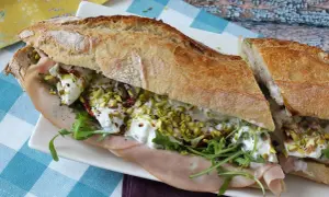 Un sandwich baguette garni de jambon, roquette, tomates séchées, pistaches concassées et burrata crémeuse.