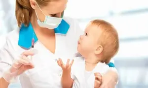 Pielęgniarka z maską i rękawiczkami trzyma strzykawkę z różowym płynem, przygotowując się do podania szczepionki 5w1 czy 6w1 dziecku.