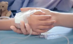 Dłoń dorosłego obejmuje dłoń dziecka z wenflonem. W tle pluszowy miś. Dokumentacja do orzeczenia o niepełnosprawności i zasiłek pielęgnacyjny.
