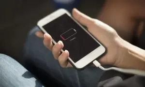 Dłoń trzyma smartfon z ikoną niskiego poziomu baterii i napisem "Charging". Wtyczka kabla ładującego jest podłączona. Oszczędzanie baterii jest kluczowe.