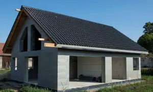 Dom w stanie surowym otwartym, z betonowych pustaków, z czarnym dachem i otwartymi otworami okiennymi i drzwiowymi.