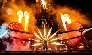 Płomienie buchają na koncercie Rammstein, tworząc piekielną atmosferę. Wokalista stoi w centrum, otoczony ogniem.