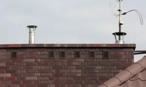 Zakończenie komina wentylacyjnego z metalową nasadką i kratkami wentylacyjnymi w ceglanej ścianie. Obok widoczna antena telewizyjna.