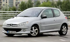 Srebrny Peugeot 206 z widocznymi przednimi żarówkami, zaparkowany na mokrym parkingu.