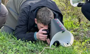 Fotograf z aparatem i dyfuzorem do makro pochyla się nad trawą, by uchwycić drobne detale natury.