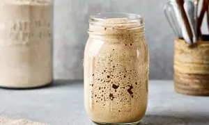 Ein Glas mit sprudelndem Sauerteig, bereit zum Backen. Im Hintergrund ein weiteres Glas mit Mehl und Küchenutensilien.