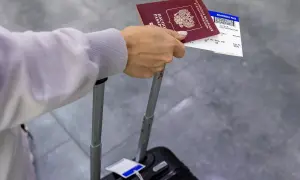 Dłoń trzyma paszport i kartę pokładową, która jest biletem na samolot. Obok walizka.