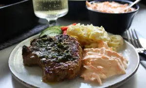 En tallrik med saftiga kotletter i ugn, potatisgratäng och en krämig morotsröra. Ett glas vitt vin står bredvid.