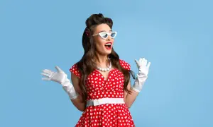 Kobieta w stylu pin-up w czerwonej sukience w białe grochy, białych rękawiczkach i okularach, z uniesionymi rękami.