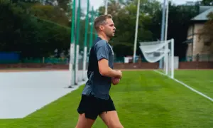 Mężczyzna w sportowym stroju biegnie po bieżni. W tle widać boisko z bramką i drzewa. bc1 bieganie.