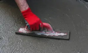 Dłoń w czerwonej rękawicy wygładza świeży beton kielnią. To kluczowy etap prac, gdzie dba się o równomierne rozłożenie materiału, co jest ważne dla późniejszego zbrojenia rozproszonego.