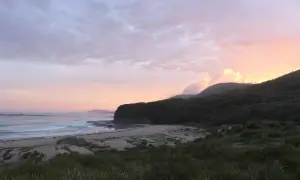 Zachód słońca nad dzikim wybrzeżem, z falami rozbijającymi się o piaszczystą plażę i zalesione wzgórza w tle. Widok na east coast o wschodzie słońca.