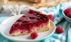 Ein saftiges Stück Himbeerkuchen mit gefrorenen Himbeeren, glasiert und auf einem Teller mit frischen Beeren daneben.