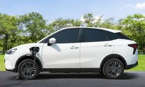 Auto elettrica ibrida bianca in carica, con cavo collegato al lato sinistro. Sfondo di alberi verdi e cielo azzurro.