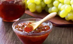 Un cuenco de cristal con mermelada de uvas casera, una cuchara de madera y un racimo de uvas verdes al fondo.