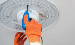 Ręka w pomarańczowej rękawicy przykręca śrubokrętem lampę sufitową LED. Dowiedz się, jak zrobić oświetlenie.