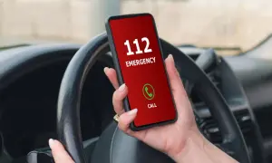 Mano sujetando un teléfono con pantalla roja que muestra "112 EMERGENCY" y un botón de llamada verde. El volante del coche está visible.